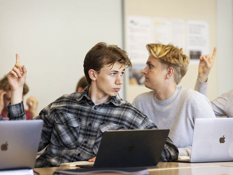 To elever arbejder på computere i et klasselokale. Den ene rækker hånden op
