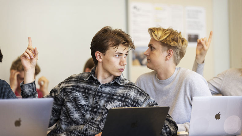To elever arbejder på computere i et klasselokale. Den ene rækker hånden op