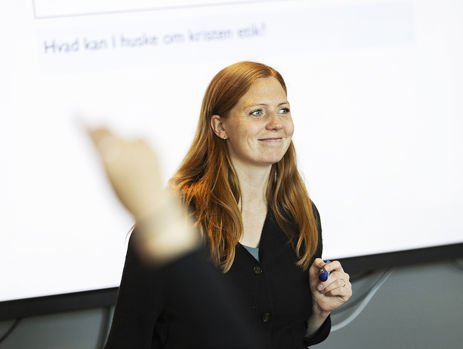 En gymnasielærer står foran et smartboard og smiler, mens en elev rækker hånden op