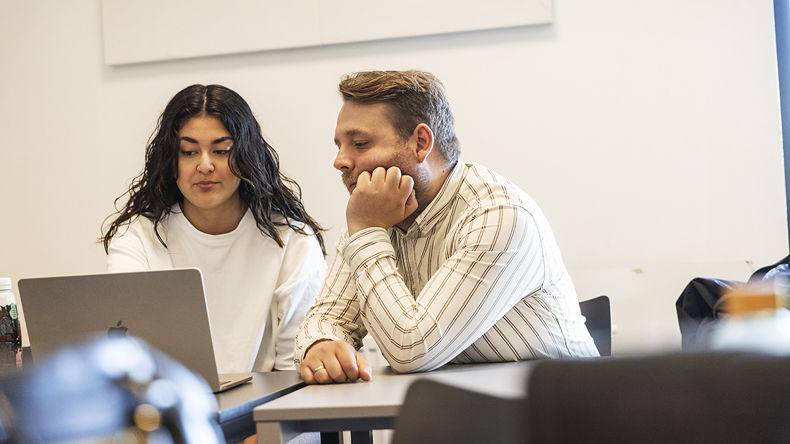 To elever sidder ved et bord og kigger sammen på en computer