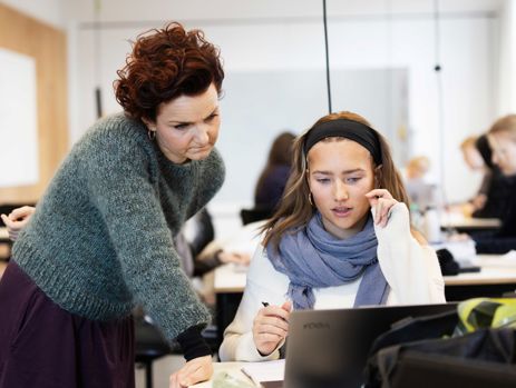 Gymnasielærer hjælper elev med arbejde på computer i klasselokalet.